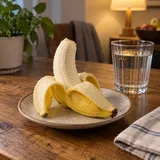 Częściowo obrany banan na ceramicznym talerzu obok szklanki wody w wieczornym świetle.