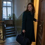 Lena z granatową torbą domyka drzwi mieszkania i zatrzymuje się na klatce schodowej o świcie.